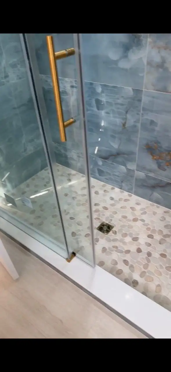 Walk-in shower with a brass-handled glass door, blue-gray marble-look wall tile and a pebble mosaic floor